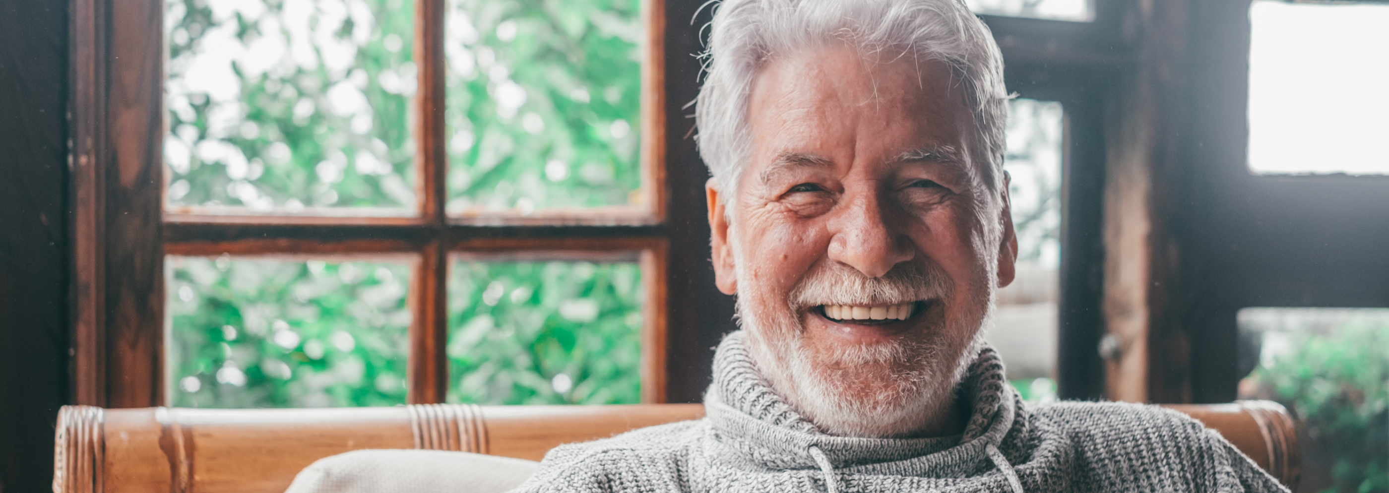 Elderly man with gray hair and beard smiling warmly in a cozy room. He wears a gray sweater, with large windows and greenery in the background.