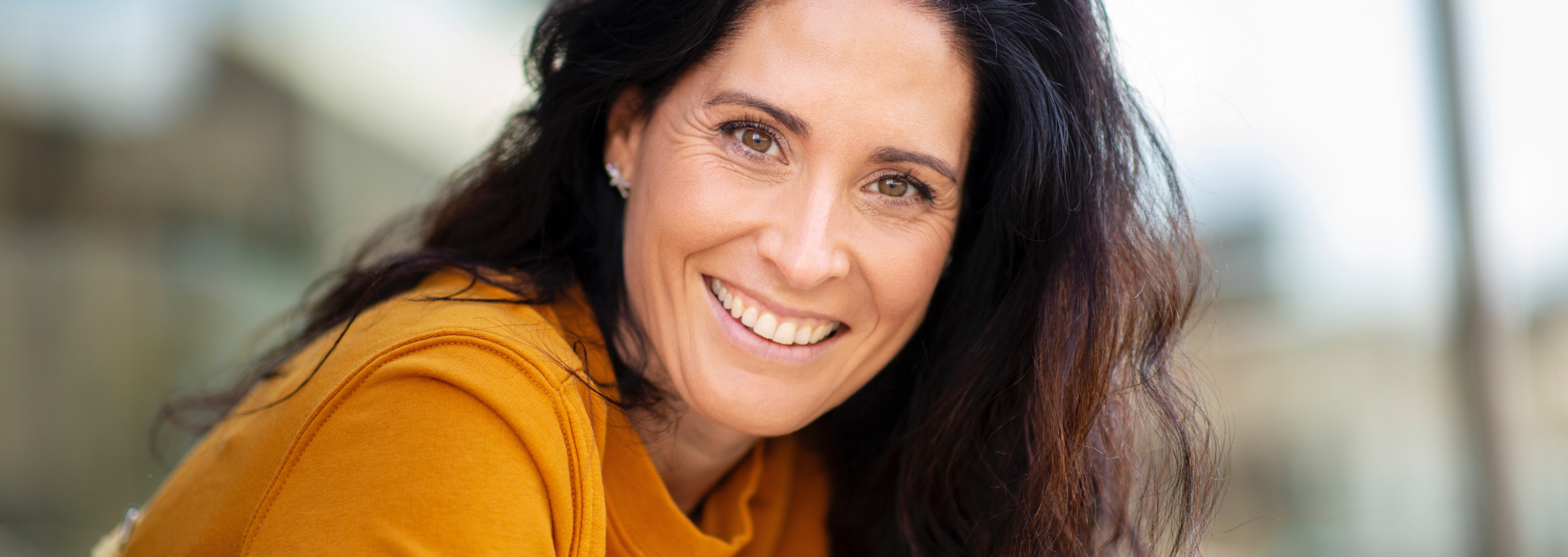 Smiling woman with long dark hair in a mustard yellow top, leaning slightly forward. Background is softly blurred. The mood is cheerful and welcoming.