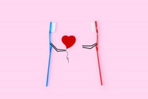 A blue toothbrush handing a red heart balloon to a red toothbrush